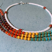 Load image into Gallery viewer, A multi-strand beaded necklace with a white cord base and rows of colorful beads in red, orange, yellow, teal, and purple, accented with gold spacer beads, displayed on a textured gray surface.