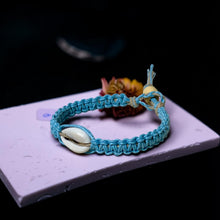 Load image into Gallery viewer, Blue braided bracelet with a shell on a purple surface