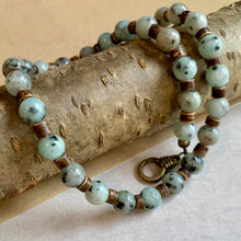 Load image into Gallery viewer, A handcrafted necklace with semi-translucent blue sesame jasper gemstone beads, antiqued brass beads, and a brass lobster clasp, displayed on a wooden branch.