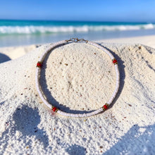 Load image into Gallery viewer, A beaded anklet with a textured white body and small red beads, displayed on a sandy beach with the ocean in the background.