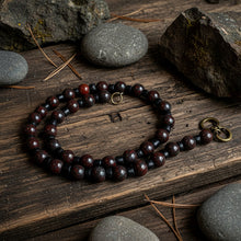 Load image into Gallery viewer, A beaded necklace made of dark reddish-brown polished round beads separated by small black beads, with a small gold-toned ring clasp and a brass lobster clasp, photographed on a light surface with a bark prop at the top.