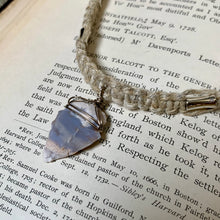 Load image into Gallery viewer, A handmade beaded necklace with a natural blue-gray pointed stone pendant wrapped in silver wire, resting on an open book with printed text.