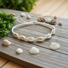 Load image into Gallery viewer, A white hemp choker necklace with cowrie shell accents, displayed on a greenish surface with some shells separately placed around the necklace.