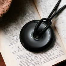 Load image into Gallery viewer, A black onyx donut pendant on a leather cord choker necklace, displayed on an open book.
