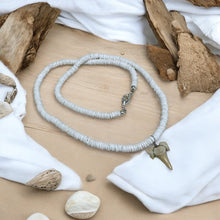 Load image into Gallery viewer, A handcrafted tribal necklace with white puka shell beads and a shark tooth pendant, displayed on a wooden surface with driftwood and shells around it.