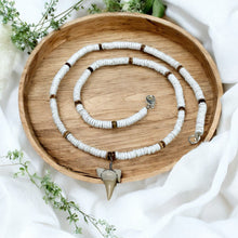 Load image into Gallery viewer, A handcrafted tribal necklace with white litub puka shell beads and a silver-plated shark tooth pendant, displayed on a wooden tray with a white fabric background.