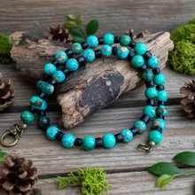 Load image into Gallery viewer, A handcrafted magnesite necklace with teal blue gemstone beads, black coconut shell beads, and brass lobster clasp, displayed on a wooden background.