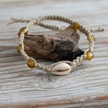 Load image into Gallery viewer, A beige hemp necklace with large cowrie shell and golden glass beads, displayed on a wooden surface.