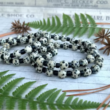 Load image into Gallery viewer, A handcrafted gemstone necklace with Dalmatian Jasper beads, black coconut shell beads, and brass lobster clasp, displayed in front of a rustic wooden background.