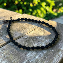 Load image into Gallery viewer, A black macrame beaded bracelet with a tassel, displayed on a weathered wooden surface with a blurred green garden background.
