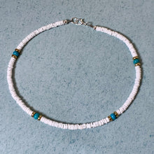 Load image into Gallery viewer, A beaded necklace made of small white disc-like beads with four turquoise-blue accent beads and small gold spacer rings, laid flat on a textured light blue-gray surface.
