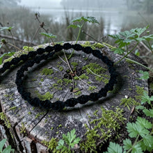 Load image into Gallery viewer, A black braided cord or rope accessory arranged in a loose circle on a mossy tree stump outdoors, with blurred greenery and water in the background.
