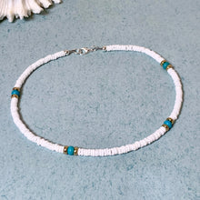 Load image into Gallery viewer, A beaded necklace made of small white disc-like beads with four turquoise-blue spacer beads, each surrounded by gold-toned metal caps, displayed on a textured light blue surface.
