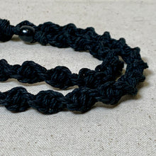 Load image into Gallery viewer, A close-up of a black braided cord necklace or bracelet with a glossy black bead, laid on a textured beige fabric background.
