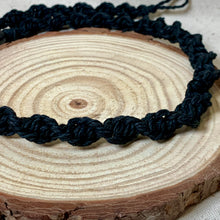 Load image into Gallery viewer, A close-up of a black braided cord  necklace draped over a round natural wood slice coaster.
