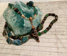 Load image into Gallery viewer, A beaded necklace with polished teal stones, dark black beads, green beads, and copper wire-wrapped accents, displayed on a large green crystal cluster over a light wood background.
