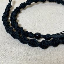 Load image into Gallery viewer, A close-up of a black macrame-style cord bracelet or necklace with a glossy black bead and tassel, laid on a light beige textured fabric background.

