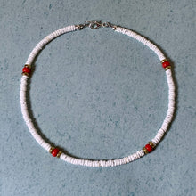 Load image into Gallery viewer, A beaded necklace made of small white disc beads with four red accent beads and small metallic spacer details, laid flat on a textured light blue-gray surface.
