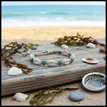Load image into Gallery viewer, A handmade macrame bracelet featuring white cowrie shells and teal blue beads, displayed on a textured dark wood branch over a beige woven fabric background.
