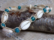 Load image into Gallery viewer, A handmade bohemian-style bracelet featuring white cowrie shells, turquoise blue beads, and natural tan cord, displayed on a textured gray bark branch over a beige woven fabric background.
