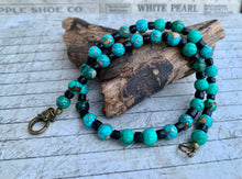 Load image into Gallery viewer, A beaded necklace with turquoise and black beads, featuring a brass lobster clasp, displayed on a piece of driftwood over a vintage-style printed background.
